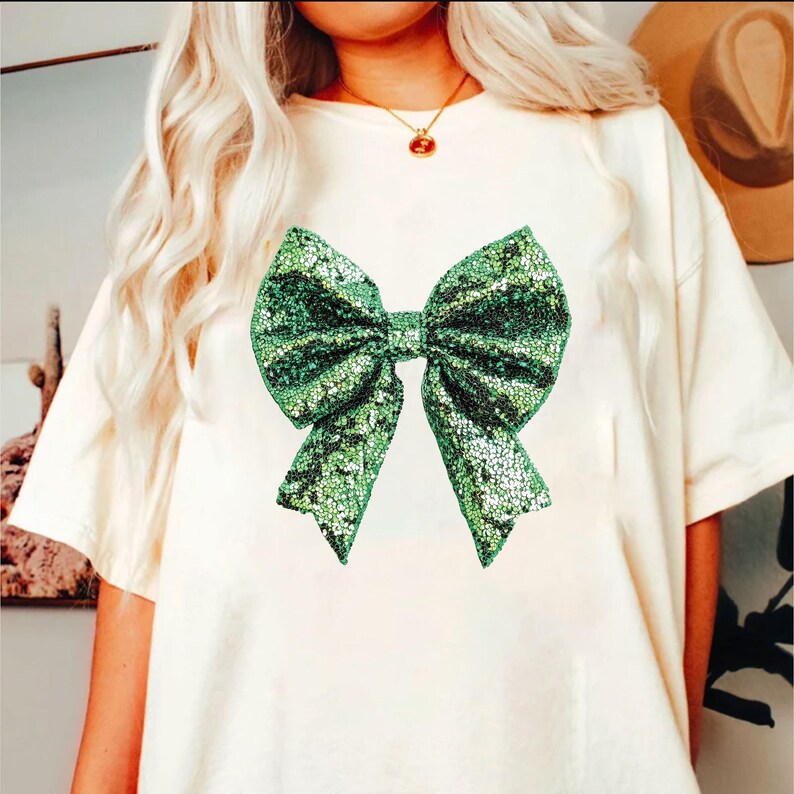 Puede incluir: Camiseta color crema con un gran dise&ntilde;o de lazo verde brillante. El lazo, hecho de lentejuelas, es el elemento central del dise&ntilde;o. La camiseta tiene mangas cortas y un corte holgado.