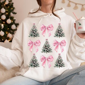 May include: White hoodie with a repeating pattern of Christmas trees and pink bows. The trees are green with white detailing, and the bows are a soft pink. The design is repeated across the front. A casual, festive garment for the holiday season.