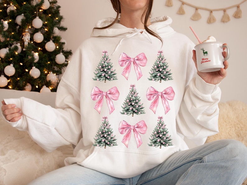 May include: White hoodie with a repeating pattern of Christmas trees and pink bows. The trees are green with white detailing, and the bows are a soft pink. The design is repeated across the front. A casual, festive garment for the holiday season.