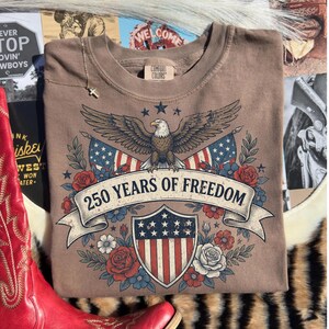 May include: A brown Comfort Colors crew neck t-shirt featuring an eagle, American flags, and the text "250 YEARS OF FREEDOM." The design includes a shield with stars and stripes, surrounded by red and blue flowers. A gold necklace with a cross is visible.