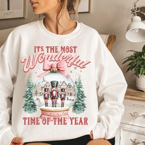 May include: White sweatshirt with a pink and red graphic design featuring a snow globe with nutcracker figurines and the text "It's the most Wonderful Time of the Year".