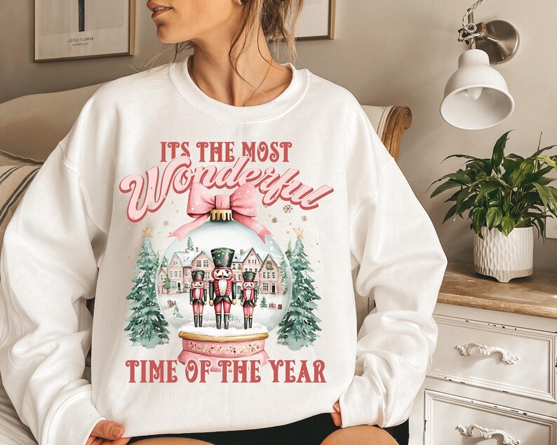 May include: White sweatshirt with a pink and red graphic design featuring a snow globe with nutcracker figurines and the text "It's the most Wonderful Time of the Year".