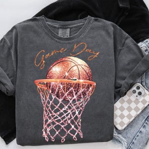 May include: A charcoal grey t-shirt with the text "Game Day" in gold glitter lettering. A glittery orange basketball is shown going through a pink basketball hoop.