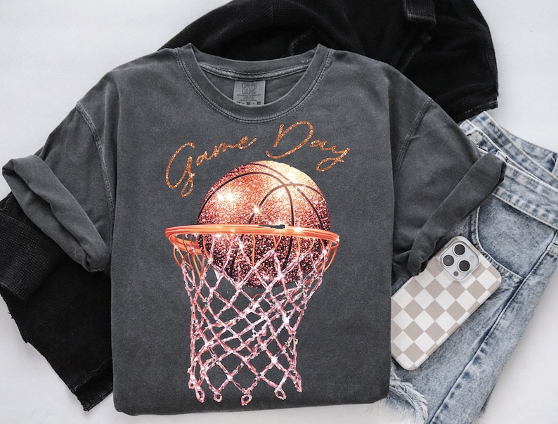May include: A charcoal grey t-shirt with the text "Game Day" in gold glitter lettering. A glittery orange basketball is shown going through a pink basketball hoop.
