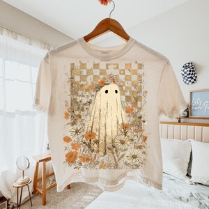 May include: Cream-colored t-shirt featuring a ghost design. The ghost has two black eyes and a draped sheet appearance. The shirt's graphic includes a floral arrangement with daisies and a checkered background in shades of yellow and brown.