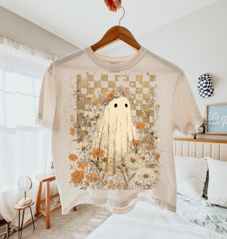 May include: Cream-colored t-shirt featuring a ghost design. The ghost has two black eyes and a draped sheet appearance. The shirt's graphic includes a floral arrangement with daisies and a checkered background in shades of yellow and brown.
