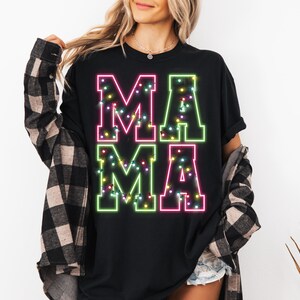 May include: Black t-shirt with the word "MAMA" in large, neon-style letters. The letters are outlined in green and pink, with a string of colorful lights inside each letter. The shirt is a casual, everyday wear item.
