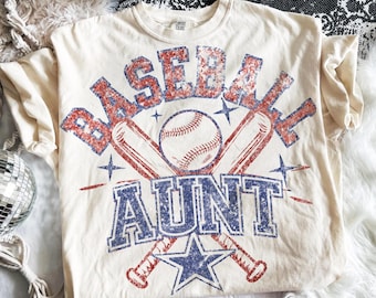 May include: Cream-colored t-shirt with a baseball-themed graphic. The design features the words "BASEBALL AUNT" in distressed red and blue lettering, along with a baseball, crossed bats, and stars. The shirt is made of a soft material.