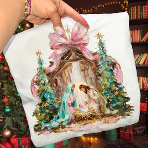 May include: White pillow featuring a Nativity scene with a pink bow, gold accents, and two Christmas trees. The artwork depicts Mary, Joseph, and baby Jesus in a manger. The pillow is held up by a hand.