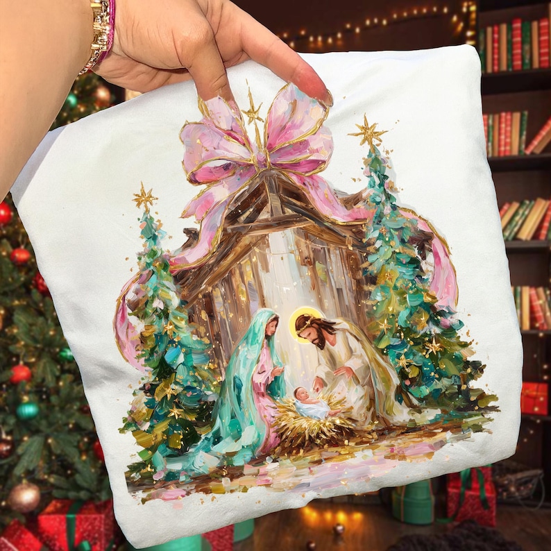 May include: White pillow featuring a Nativity scene with a pink bow, gold accents, and two Christmas trees. The artwork depicts Mary, Joseph, and baby Jesus in a manger. The pillow is held up by a hand.