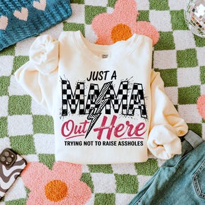 Puede incluir: Una sudadera de color crema con un gr&aacute;fico a cuadros en blanco y negro que dice "Just a Mama Out Here Trying Not to Raise Assholes".