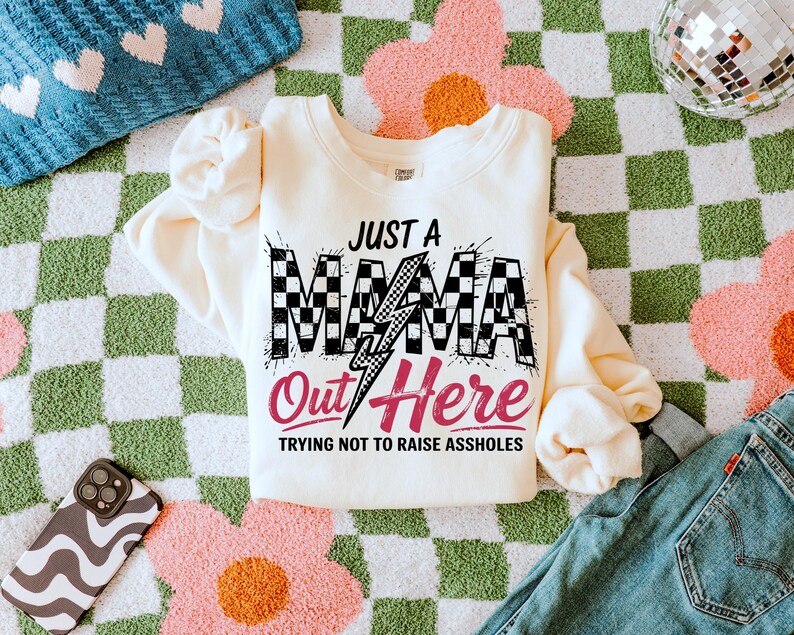 Puede incluir: Una sudadera de color crema con un gr&aacute;fico a cuadros en blanco y negro que dice "Just a Mama Out Here Trying Not to Raise Assholes".