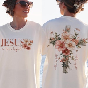 May include: White t-shirt with the text "JESUS is the light" on the front and a floral cross design on the back. The cross is surrounded by pink and white flowers and the text "walk by faith" and "truly, truly I say to you" are written on the cross.