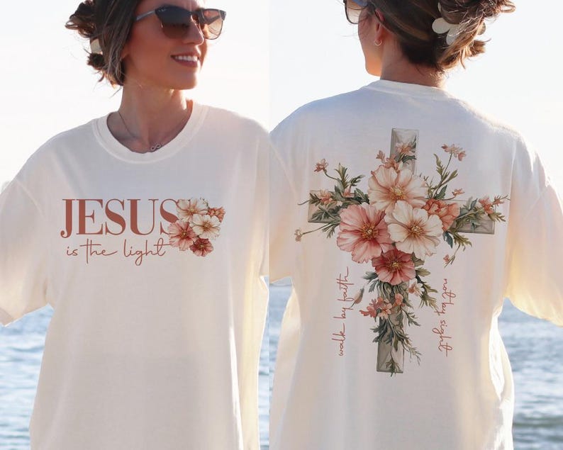 May include: White t-shirt with the text "JESUS is the light" on the front and a floral cross design on the back. The cross is surrounded by pink and white flowers and the text "walk by faith" and "truly, truly I say to you" are written on the cross.