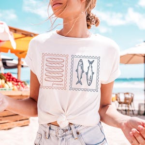 May include: White t-shirt featuring two postage stamp designs. One stamp shows six brown bread loaves with the word "forever" below. The other stamp displays two fish. The shirt is tied at the waist.