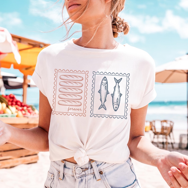 May include: White t-shirt featuring two postage stamp designs. One stamp shows six brown bread loaves with the word "forever" below. The other stamp displays two fish. The shirt is tied at the waist.