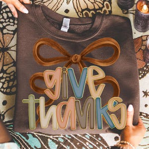 May include: A brown sweatshirt with the words "GIVE THANKS" in colorful, outlined letters. A large brown bow graphic is above the text. The sweatshirt is being held up by a person.