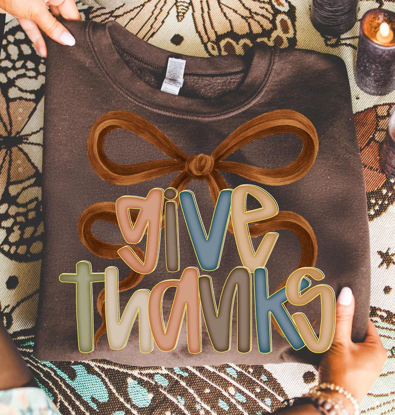 May include: A brown sweatshirt with the words "GIVE THANKS" in colorful, outlined letters. A large brown bow graphic is above the text. The sweatshirt is being held up by a person.