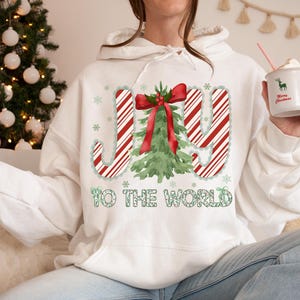 Puede incluir: Sudadera con capucha blanca con un dise&ntilde;o navide&ntilde;o. El dise&ntilde;o presenta la palabra "JOY" en rayas rojas y blancas de bast&oacute;n de caramelo, un &aacute;rbol de Navidad verde y la frase "TO THE WORLD" en verde. Se sostiene una taza con un ciervo y "Merry Christmas".