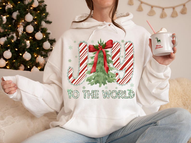 Puede incluir: Sudadera con capucha blanca con un dise&ntilde;o navide&ntilde;o. El dise&ntilde;o presenta la palabra "JOY" en rayas rojas y blancas de bast&oacute;n de caramelo, un &aacute;rbol de Navidad verde y la frase "TO THE WORLD" en verde. Se sostiene una taza con un ciervo y "Merry Christmas".