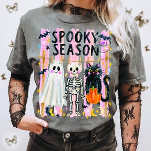 K&ouml;nnte beinhalten: Ein anthrazitfarbenes Kurzarm-T-Shirt mit einem Grafikdruck und dem Text SPOOKY SEASON. Das Motiv zeigt ein wei&szlig;es Gespenst, ein Skelett und eine schwarze Katze, die einen orangefarbenen K&uuml;rbis h&auml;lt, alles an einer W&auml;scheleine &uuml;ber rosa Streifen.