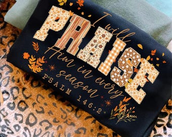 May include: Black sweatshirt featuring the word "PRAISE" and the phrase "I will praise Him in every season" in a fall design. The letters are decorated with floral and plaid patterns, and autumn leaves. Psalm 146:2 is also printed on the shirt.