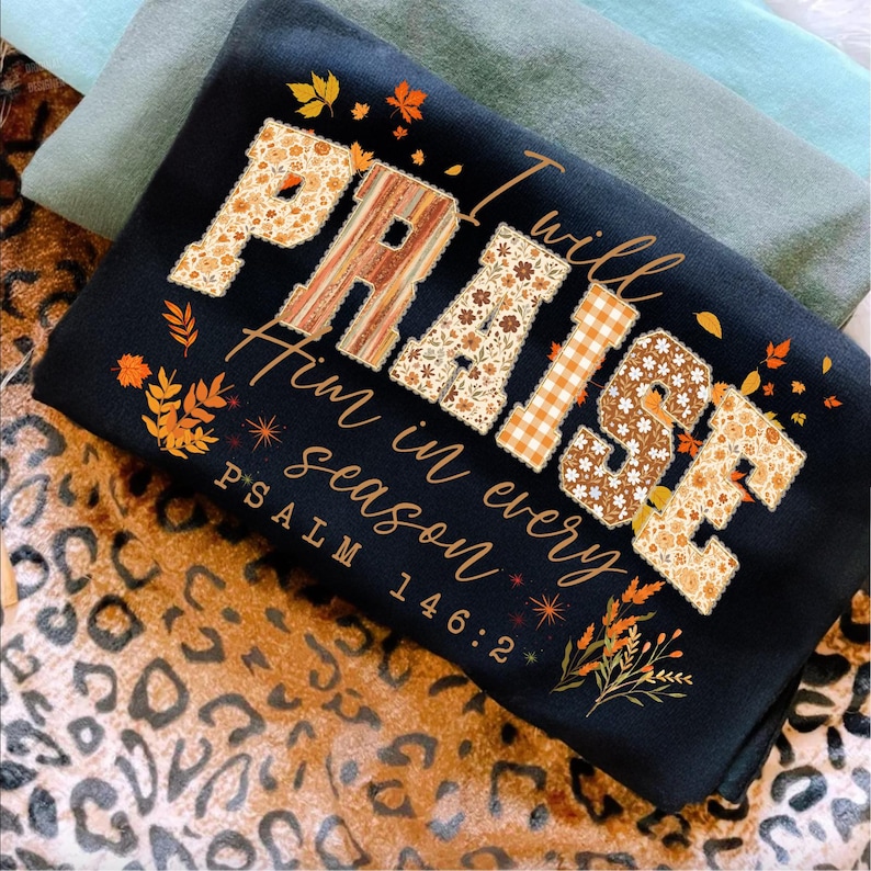 May include: Black sweatshirt featuring the word "PRAISE" and the phrase "I will praise Him in every season" in a fall design. The letters are decorated with floral and plaid patterns, and autumn leaves. Psalm 146:2 is also printed on the shirt.
