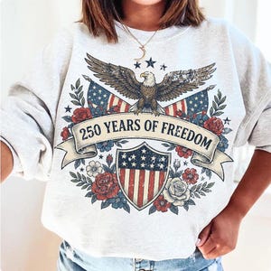 May include: Light gray sweatshirt with a patriotic design. Features an eagle, American flags, and the text "250 YEARS OF FREEDOM" on a banner. Decorated with red, white, and blue floral elements.