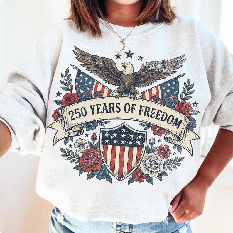 May include: Light gray sweatshirt with a patriotic design. Features an eagle, American flags, and the text "250 YEARS OF FREEDOM" on a banner. Decorated with red, white, and blue floral elements.