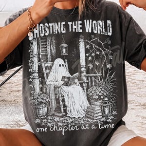 May include: A charcoal gray t-shirt featuring a white graphic print of a ghost sitting in a rocking chair on a porch reading a book. The text reads GHOSTING THE WORLD at the top and one chapter at a time at the bottom, with potted plants.