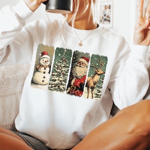 May include: White sweatshirt featuring a vintage-style Christmas design. The design includes a snowman, Christmas tree, Santa Claus, and a reindeer, all in a rectangular format. The sweatshirt is a long-sleeved crew neck.