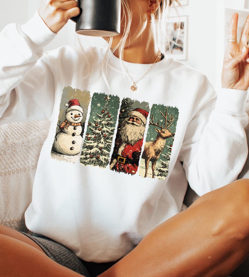 May include: White sweatshirt featuring a vintage-style Christmas design. The design includes a snowman, Christmas tree, Santa Claus, and a reindeer, all in a rectangular format. The sweatshirt is a long-sleeved crew neck.