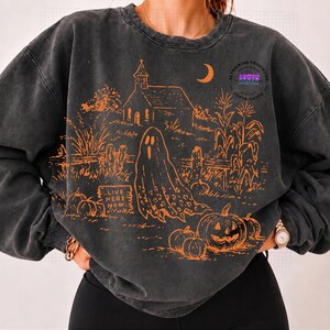 May include: A charcoal gray crewneck sweatshirt featuring an orange graphic print of a haunted scene with a church, a ghost, pumpkins, and corn stalks. The text "LIVE HERE NOW" is printed on a tombstone. The fabric has a washed, vintage look.