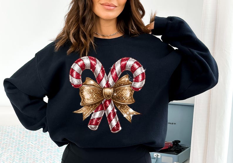 Puede incluir: Sudadera negra con un dise&ntilde;o navide&ntilde;o. El gr&aacute;fico muestra dos bastones de caramelo con rayas rojas y blancas, cruzados y atados con un lazo dorado. Una prenda festiva para la temporada navide&ntilde;a.