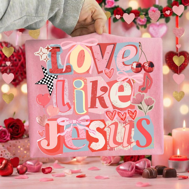 May include: Pink tote bag featuring the phrase "Love Like Jesus" in a layered, colourful design. The design incorporates hearts, stars, cherries, and musical notes. The bag is held up by a person.