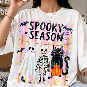 K&ouml;nnte beinhalten: Ein wei&szlig;es T-Shirt mit einem Grafikdruck, der den Text "SPOOKY SEASON" in schwarzen Buchstaben zeigt. Das Design zeigt ein Gespenst, ein Skelett und eine schwarze Katze mit einem orangefarbenen K&uuml;rbis an einer W&auml;scheleine vor rosa Streifen.