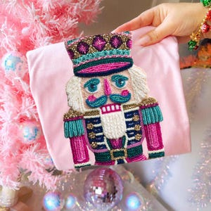 May include: A pink, embroidered Christmas stocking featuring a detailed nutcracker design. The nutcracker has a white beard, blue mustache, and a colorful uniform with gold and teal accents. The stocking is held against a pink background.