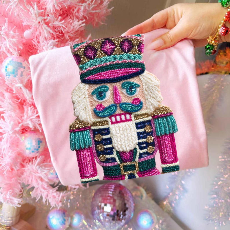 May include: A pink, embroidered Christmas stocking featuring a detailed nutcracker design. The nutcracker has a white beard, blue mustache, and a colorful uniform with gold and teal accents. The stocking is held against a pink background.