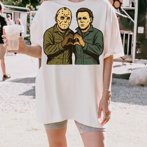 May include: Cream-colored t-shirt featuring a graphic of two horror movie characters, one wearing a hockey mask and the other with a blank expression. The characters are depicted in green and brown, forming a heart shape with their hands.