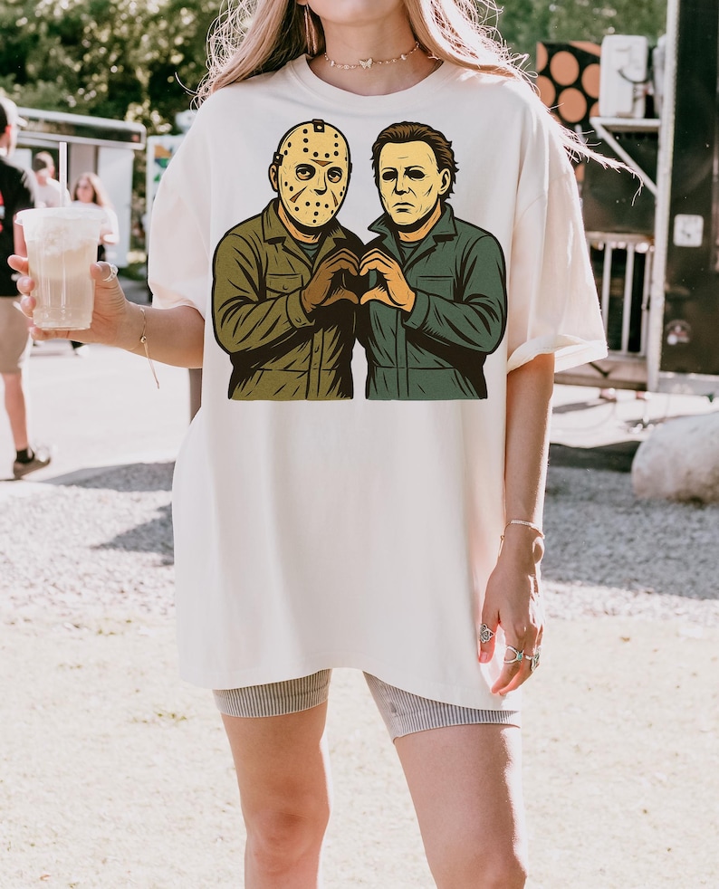 May include: Cream-colored t-shirt featuring a graphic of two horror movie characters, one wearing a hockey mask and the other with a blank expression. The characters are depicted in green and brown, forming a heart shape with their hands.