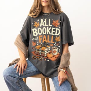 May include: Dark gray t-shirt with the text "ALL BOOKED FOR FALL" in white and orange. The graphic includes books, a pumpkin, a mug of coffee, and fall leaves. The shirt is worn with blue jeans.