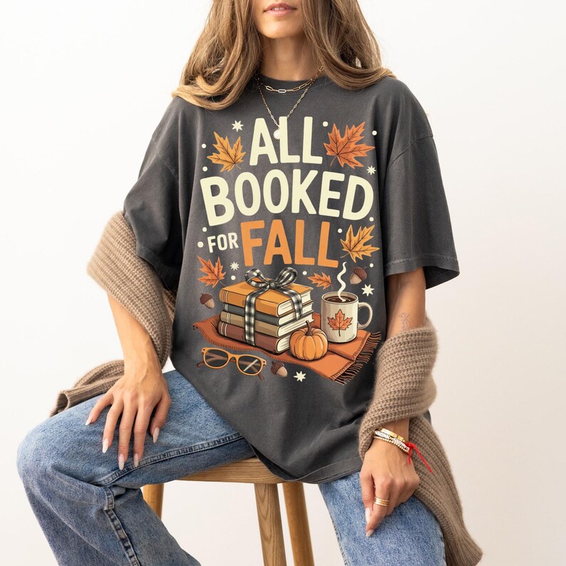 May include: Dark gray t-shirt with the text "ALL BOOKED FOR FALL" in white and orange. The graphic includes books, a pumpkin, a mug of coffee, and fall leaves. The shirt is worn with blue jeans.