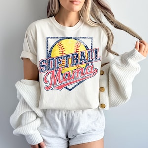 May include: Cream-colored t-shirt with a distressed graphic featuring a yellow softball, the words "SOFTBALL" in blue, and "Mama" in red. The design is set within a navy blue diamond shape.