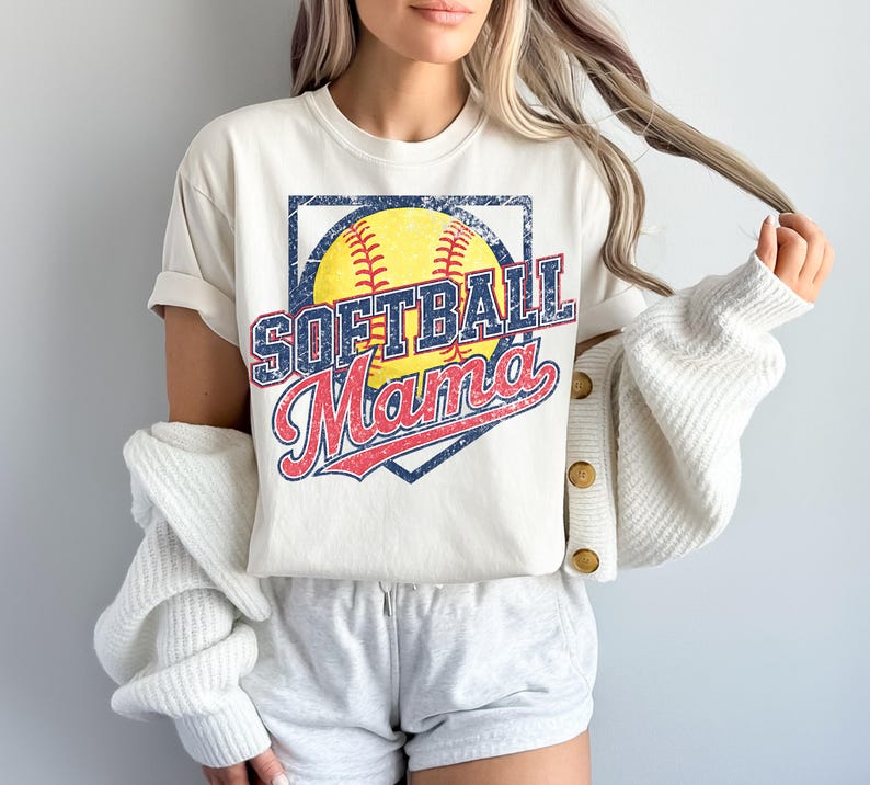 May include: Cream-colored t-shirt with a distressed graphic featuring a yellow softball, the words "SOFTBALL" in blue, and "Mama" in red. The design is set within a navy blue diamond shape.