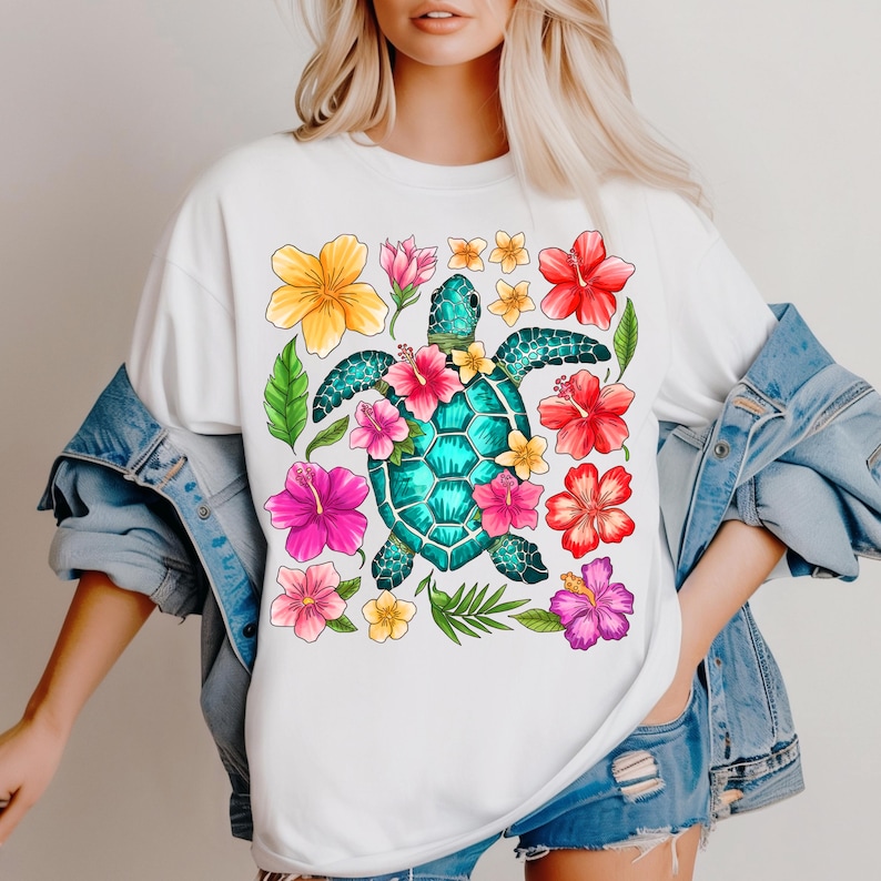 May include: White t-shirt featuring a colourful graphic of a sea turtle surrounded by tropical flowers. The turtle is turquoise blue with a green shell and is surrounded by pink, yellow, red, and purple flowers.