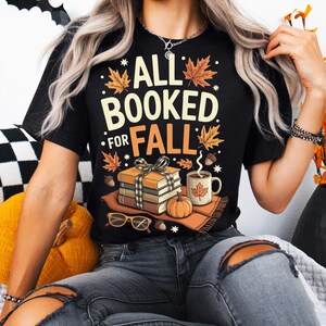 May include: Black t-shirt with the text "ALL BOOKED FOR FALL" in white and orange. The graphic includes fall leaves, a stack of books, a pumpkin, a mug of coffee, and eyeglasses. The shirt is a fall-themed design.