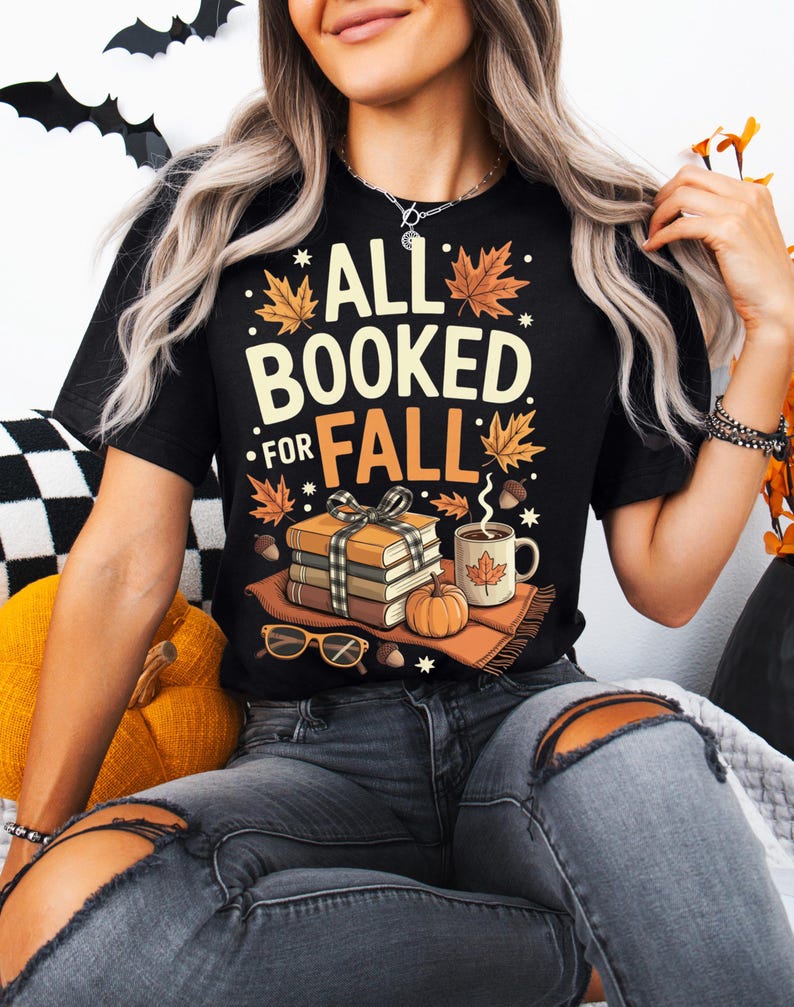 May include: Black t-shirt with the text "ALL BOOKED FOR FALL" in white and orange. The graphic includes fall leaves, a stack of books, a pumpkin, a mug of coffee, and eyeglasses. The shirt is a fall-themed design.