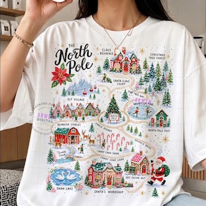 May include: A white t-shirt featuring a colorful graphic map of the North Pole. The design includes illustrations of Santa's workshop, reindeer stables, candy cane farm, and various festive buildings connected by winding paths with text labels.