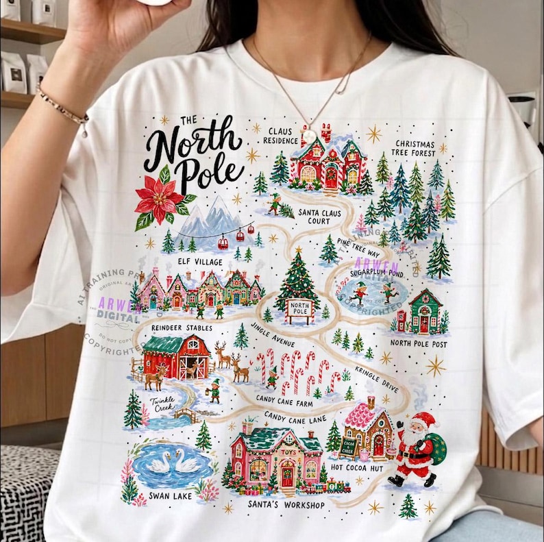 May include: A white t-shirt featuring a colorful graphic map of the North Pole. The design includes illustrations of Santa's workshop, reindeer stables, candy cane farm, and various festive buildings connected by winding paths with text labels.