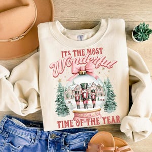May include: A cream-coloured sweatshirt with a Christmas-themed design. The design features a snow globe with three nutcracker figures inside, surrounded by houses and trees. The text "It's the most Wonderful Time of the Year" is printed below the snow globe.